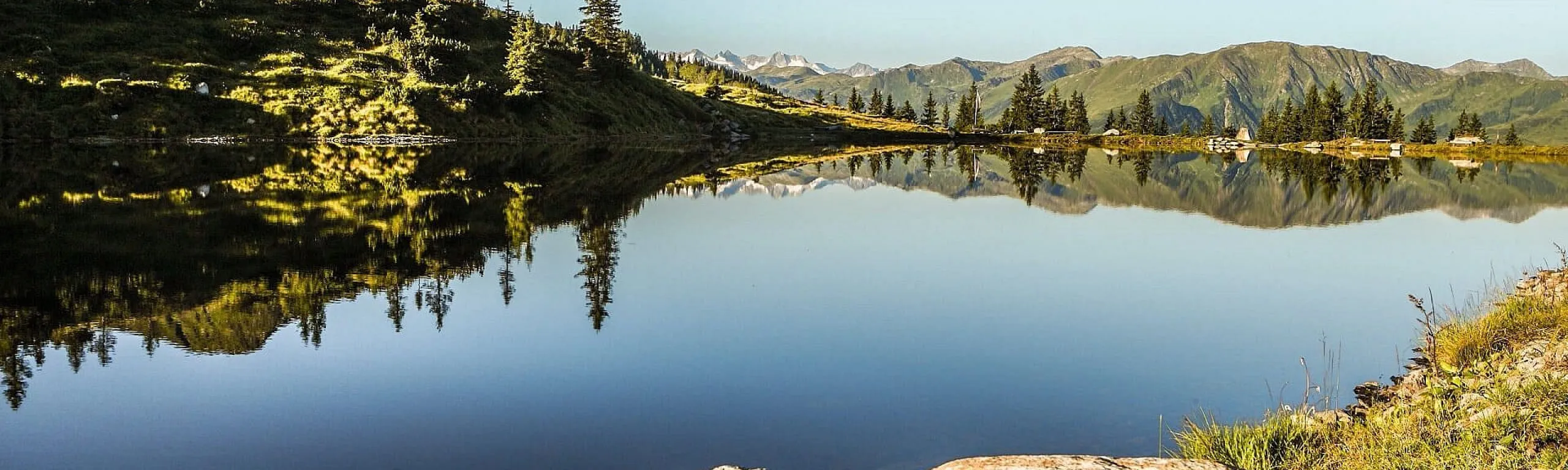 Malersicher See, umgeben von grünen Hügeln und Bäumen, spiegelt Berge im Wasser