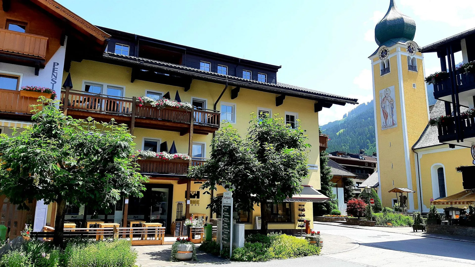 Helle Fassade eines Hotels mit Bäumen und einem Kirchturm im Hintergrund